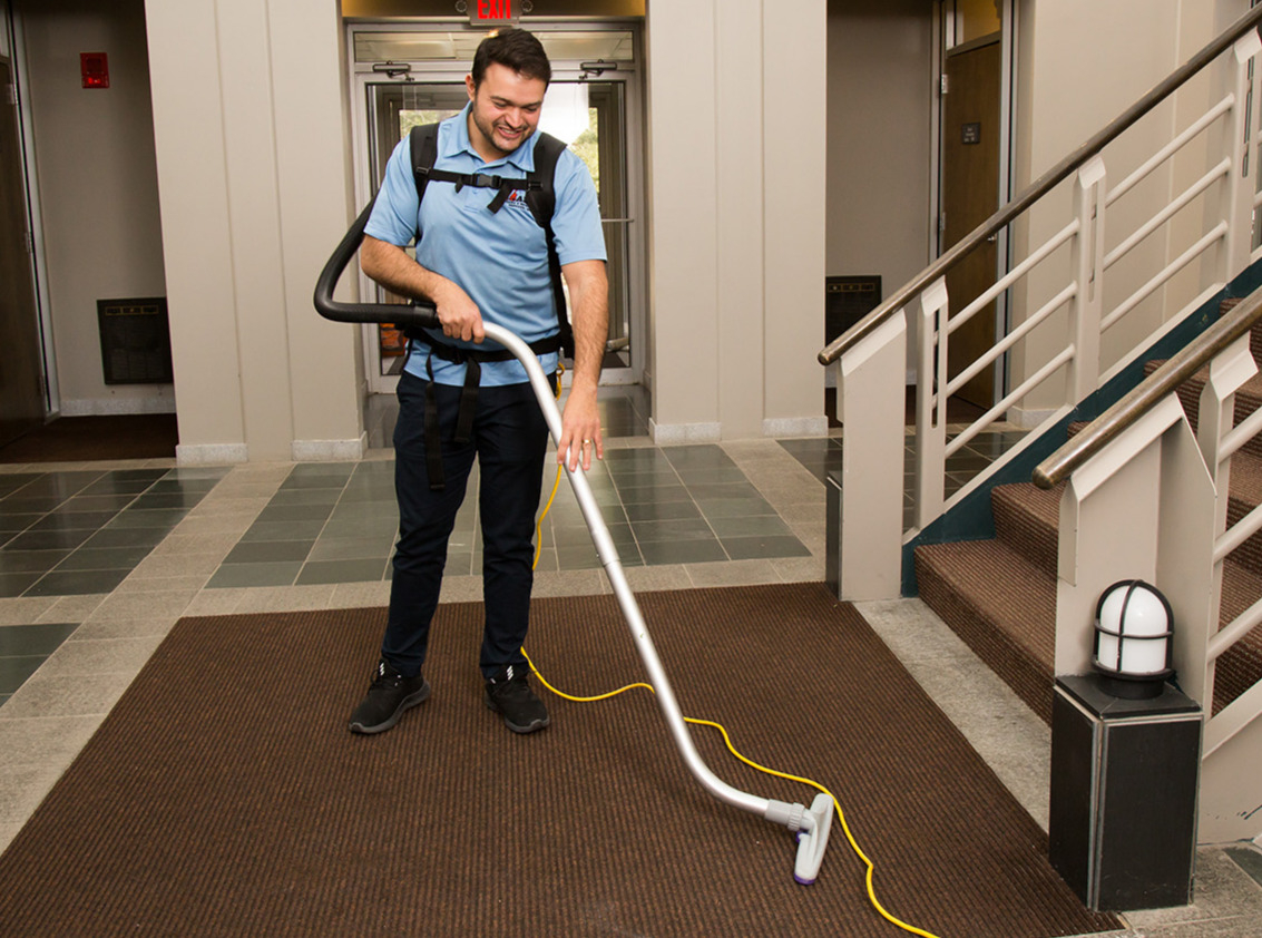 Worker using a backpack vacuum on a carpeted entrance beside a staircase.
