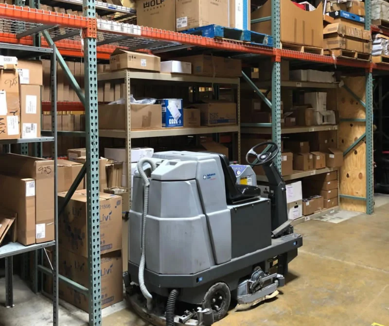 Ride-on floor cleaning machine parked between warehouse shelving